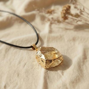 Puede incluir: Un collar con colgante dorado con una piedra de forma irregular y tallada toscamente. La piedra cuelga de un cordón negro. El colgante tiene una superficie texturizada y un brillo metálico, lo que sugiere una piedra preciosa natural.