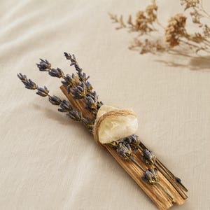 Draufsicht auf ein Palo Santo Räucherbündel mit lila Lavendelblüten und einem hellen Citrin Rohstein, sanft gebunden mit Naturkordel auf beigem Leinenstoff.