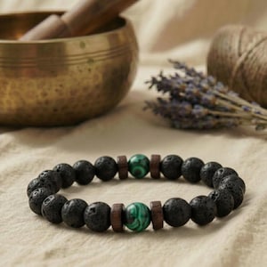 Puede incluir: Pulsera de cuentas de piedra de lava negra con detalles de malaquita verde y separadores de madera. La pulsera se muestra sobre una superficie neutra, con un cuenco tibetano de latón y ramitas de lavanda. Un accesorio natural y terroso.