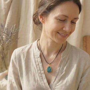 May include: A woman wearing a necklace with a teal-colored, irregularly shaped pendant. The pendant is set in a gold-colored frame and hangs from a black cord. The woman is wearing a light-colored, button-down shirt.