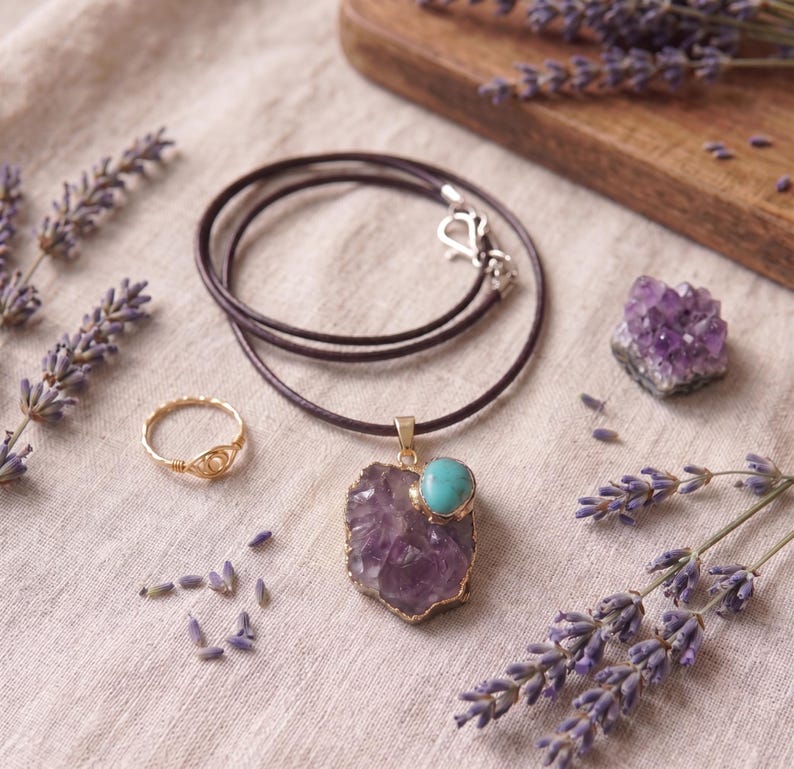 Flatlay-Ansicht der Amethyst-Talisman-Halskette mit Lederband, drapiert mit Goldring, Amethyst-Kristall und Lavendelblüten.