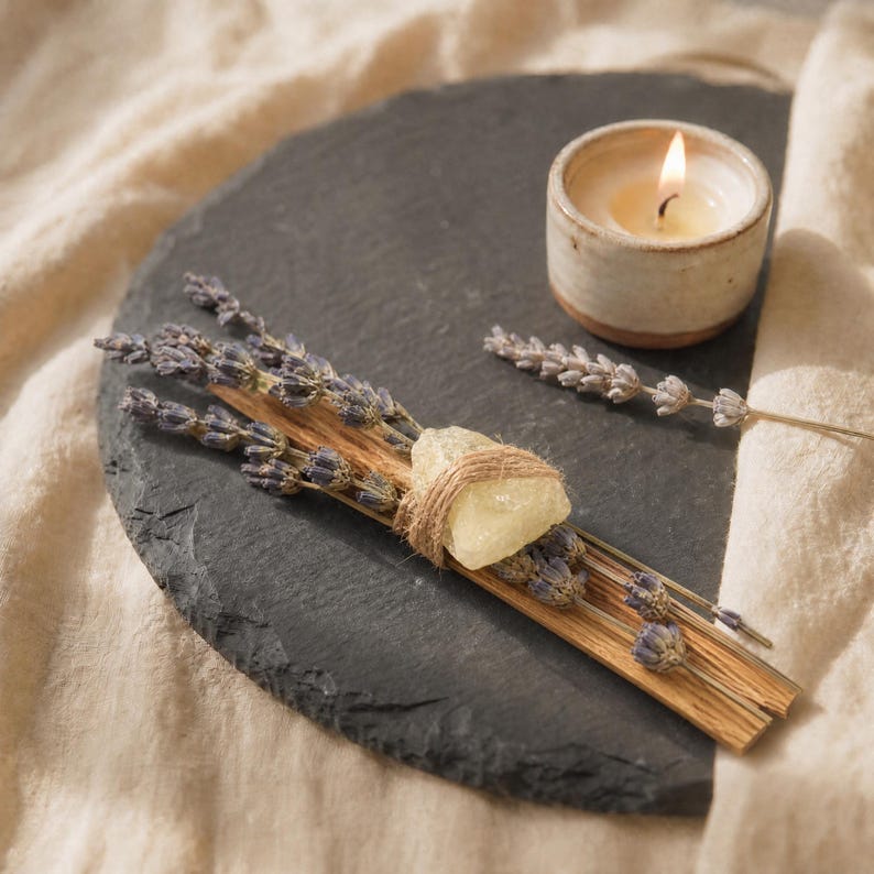 Palo Santo und Lavendel Smudge Stick mit Citrin Kristall auf einer runden schwarzen Schieferplatte, stimmungsvoll dekoriert neben einer brennenden Teelichtkerze.