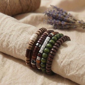 Puede incluir: Colección de seis pulseras de cuentas de madera en varios tonos de marrón, verde y crema. Algunas pulseras presentan detalles metálicos plateados. Las pulseras están dispuestas sobre una tela beige, con un ramo de lavanda seca en el fondo.