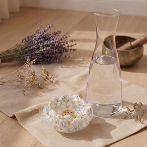 Op de afbeelding: Een heldere glazen karaf gevuld met water staat naast een wit en goud gespikkelde bloemvormige kom met een klein messing inzetstuk. Een bos gedroogde paarse lavendel en beige bloemen zijn in de buurt gerangschikt.