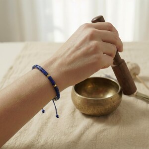 Eine geöffnete Handfläche, die das blaue Lapis Lazuli Heishi-Armband mit goldenen Zwischenperlen und Schiebeknoten präsentiert. Hintergrund: Klangschale und Lavendel.