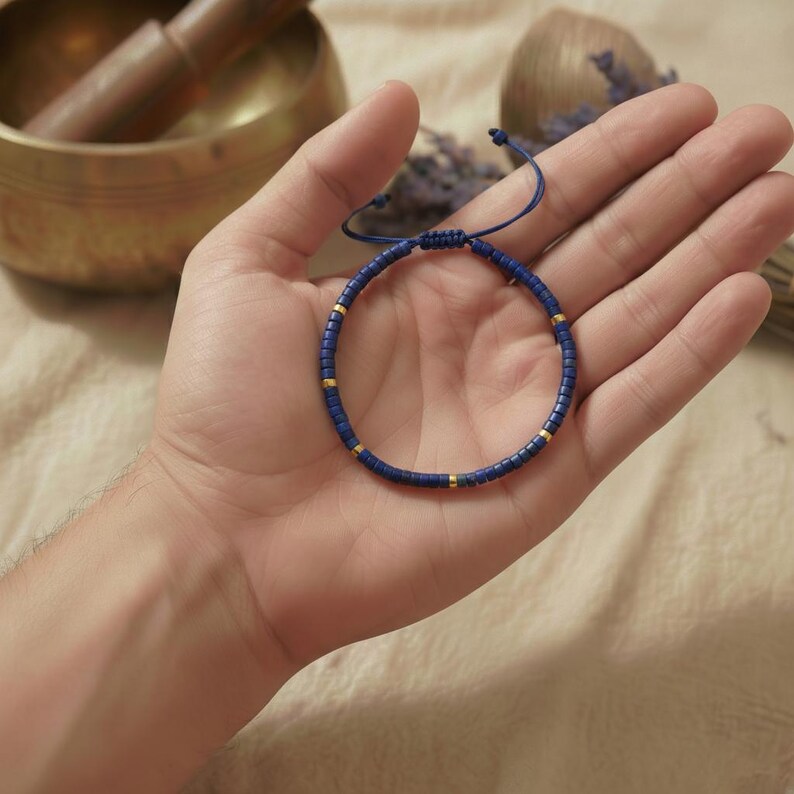 Detailaufnahme eines minimalistischen Lapis Lazuli Heishi-Perlen Armbands mit goldenen Akzenten und blauem Textil-Verstellverschluss auf beigem Leinenstoff mit Lavendel.
