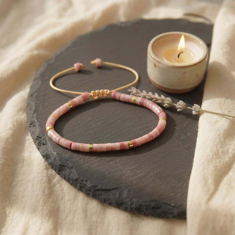 May include: A bracelet featuring small, cylindrical pink beads interspersed with thin, polished gold-tone metal spacers. The bracelet has an adjustable cord closure with two small pink beads at the ends, creating a circular shape on a dark surface.