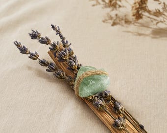 Può includere: Un mazzetto di rametti di lavanda essiccata e un bastoncino di legno marrone chiaro legati insieme con dello spago sottile. Un cristallo grezzo verde pallido è fissato al centro del mazzetto con lo stesso spago, tenendo fermi lavanda e legno.