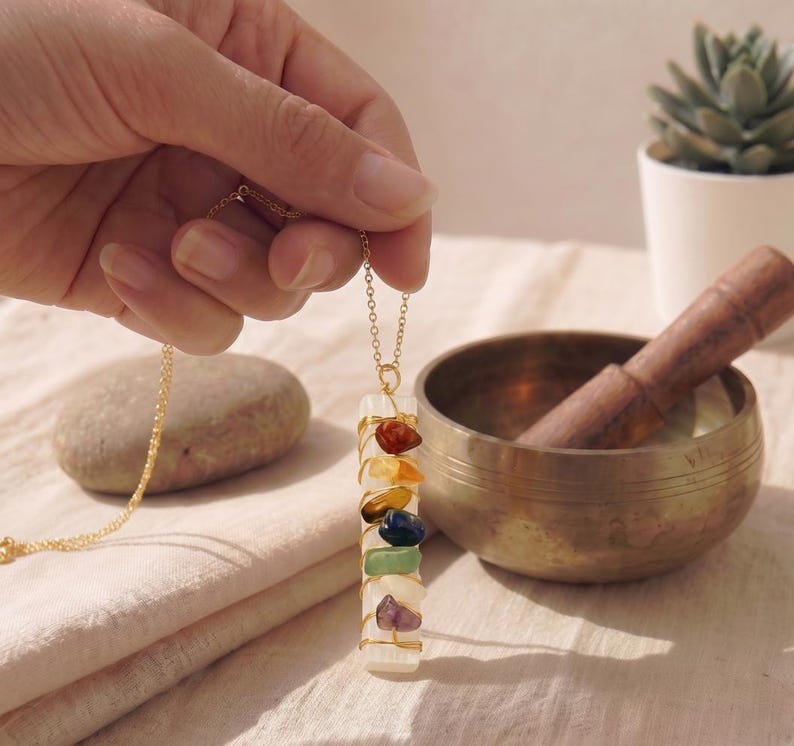 Hand hält die goldene Kette mit Selenit-Stab und sieben bunten Natursteinen im Sonnenlicht.