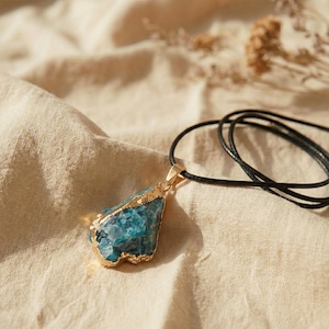 May include: A blue gemstone pendant necklace with gold-colored trim and a black cord. The gemstone has a rough, natural texture and is set in a gold-colored metal. The necklace is displayed on a beige fabric.