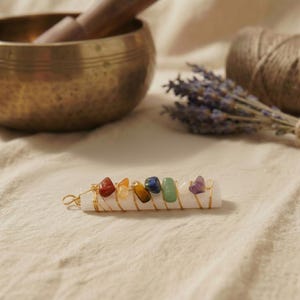 Chakra-Anhänger aus weißem Selenit mit goldfarbenem Draht und bunten Edelsteinen vor tibetischer Klangschale.