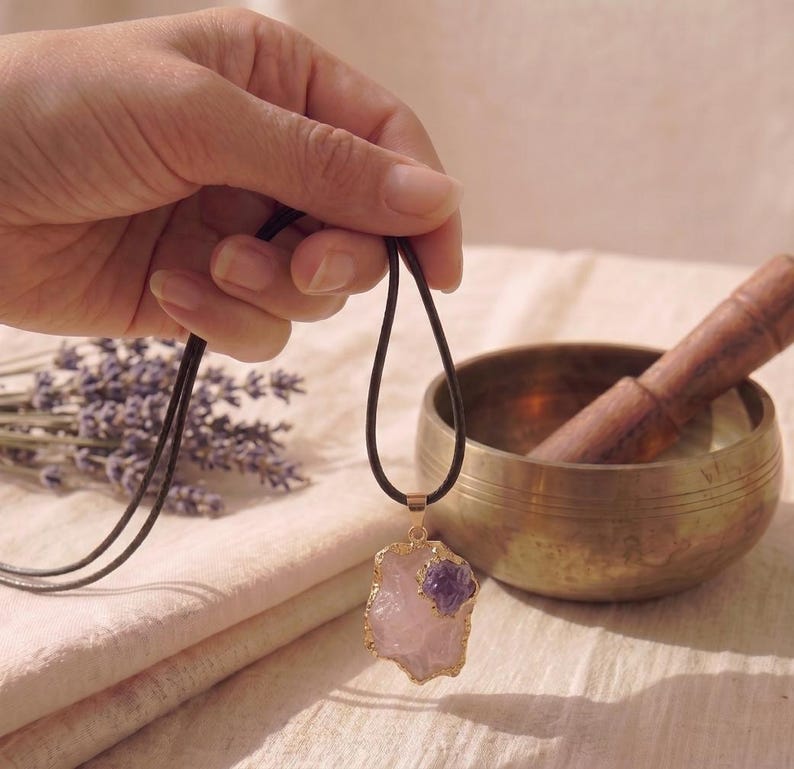 Rosenquarz Amethyst Anhänger Kette in Goldfassung gehalten in der Hand neben tibetischer Klangschale und getrocknetem Lavendel