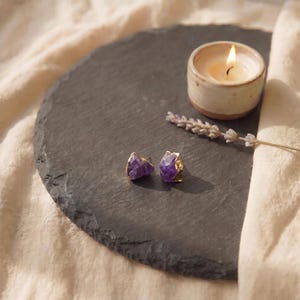 May include: A pair of stud earrings featuring raw, deep purple amethyst crystals with gold-toned metallic edges. The earrings are displayed alongside a small, lit candle in a ceramic holder and a sprig of dried lavender on a dark slate surface.
