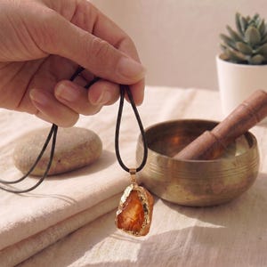 Peut inclure: Un collier pendentif composé d'une pierre précieuse orange brute et translucide avec un bord métallique doré, suspendu à un cordon noir fin. La pierre présente une forme naturelle irrégulière et est fixée par une bélière en métal doré.