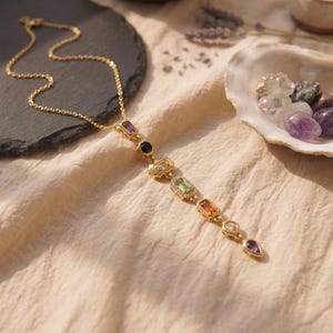 Vergoldete Chakra Y-Kette flach liegend auf Leinenstoff neben einer Muschel mit Edelsteinen und Schieferplatte.
