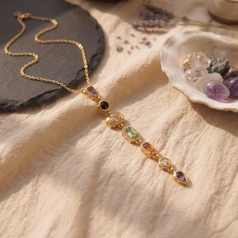 Vergoldete Chakra Y-Kette flach liegend auf Leinenstoff neben einer Muschel mit Edelsteinen und Schieferplatte.