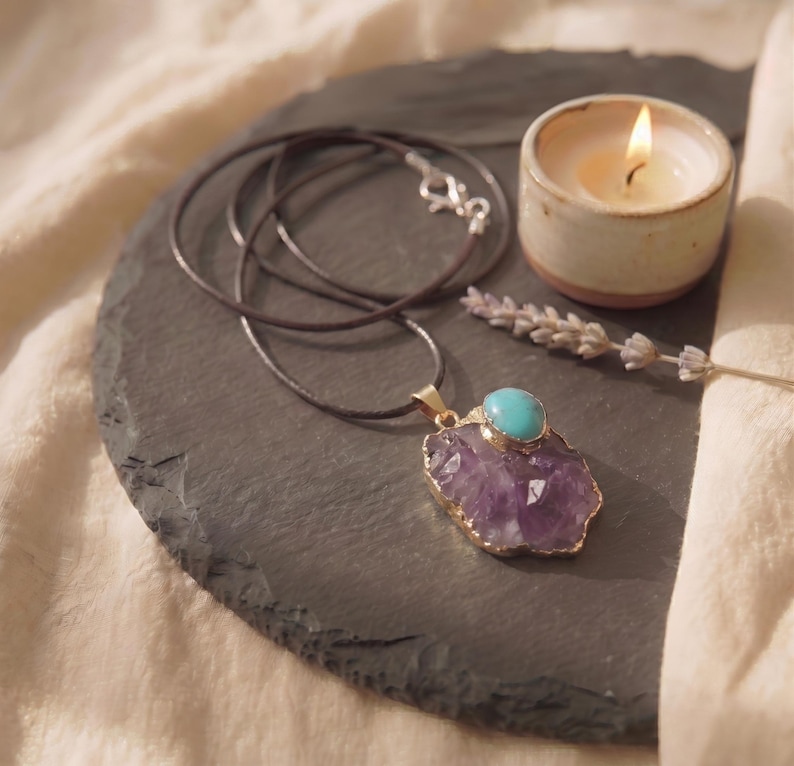 Rohstein Amethyst Kette mit gefasstem Türkis auf einer runden Schieferplatte neben einer brennenden Kerze und getrocknetem Lavendel.