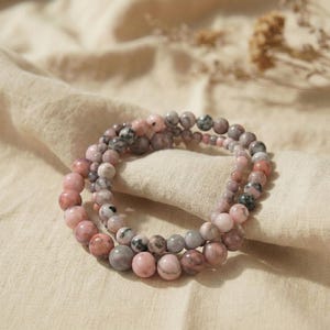 Pode incluir: Três pulseiras de contas com pedras rosa, cinza e brancas. As pulseiras são empilhadas, com contas de tamanhos variados. O fundo é um tecido bege.