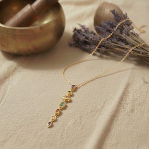 Vergoldete Chakra Kette mit bunten geometrischen Steinen auf hellem Leinenstoff neben einer tibetischen Klangschale und Lavendelzweigen.