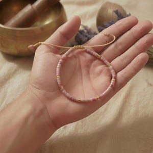 Rosa Rhodonit Perlenarmband geöffnet in einer Handfläche präsentiert mit Zugverschluss und Klangschale im Hintergrund.