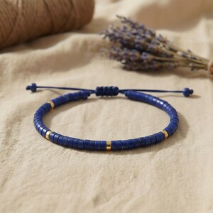 Makroaufnahme der echten Lapis Lazuli Heishi-Perlen und der goldfarbenen Metall-Akzente auf dem minimalistischen, verstellbaren Armband, liegend auf Stoff.