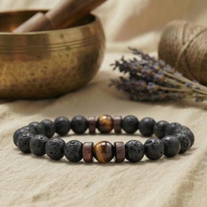 Könnte beinhalten: Ein Armband aus schwarzem Lavastein mit braunen Holzperlen und Tigeraugen-Akzenten. Das Armband ist auf einem elastischen Band aufgezogen. Das Armband wird auf einer neutralfarbenen Oberfläche präsentiert.