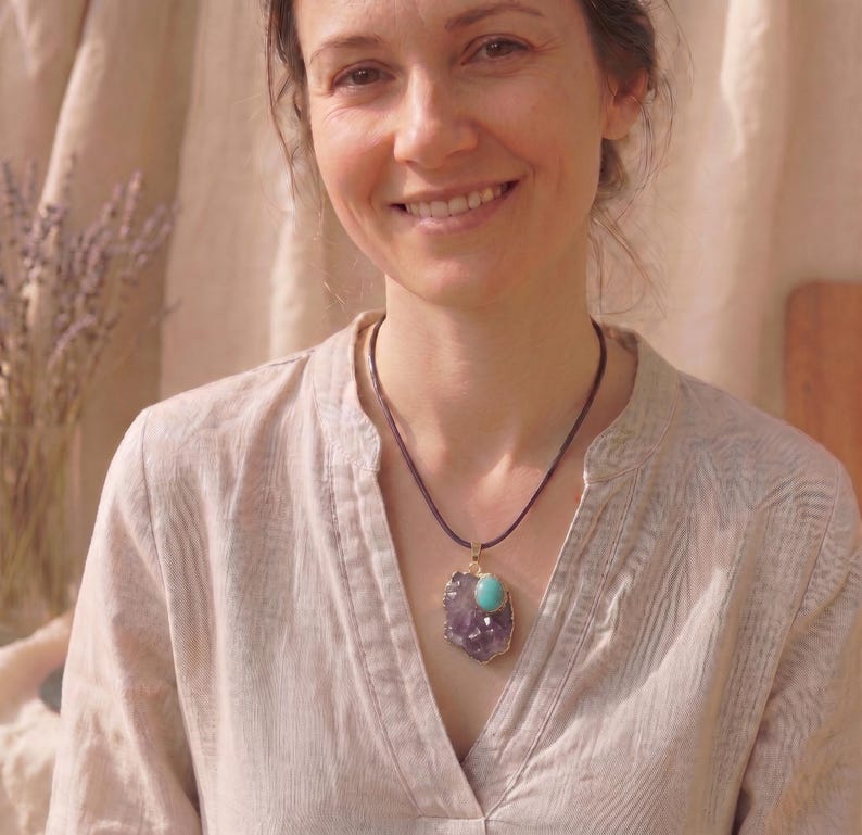 May include: A necklace featuring a dark cord and a pendant composed of a large, irregular purple amethyst crystal slice topped with a smooth, oval-shaped turquoise stone set in a gold-toned metal frame that follows the crystal's natural edge.