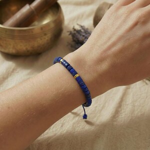 Ein zierliches, verstellbares Lapis Lazuli Armband mit Heishi-Perlen und Golddetails, getragen am Handgelenk einer Frau, im Hintergrund eine Klangschale.