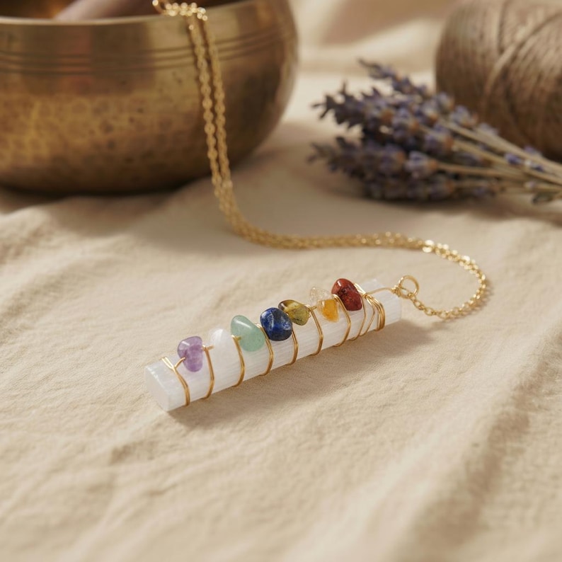 Chakra-Anhänger aus weißem Selenit mit goldfarbenem Draht und bunten Edelsteinen vor tibetischer Klangschale.