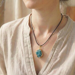 May include: A blue gemstone pendant necklace. The pendant is a rough-cut, irregular shape with a gold-colored bezel. The necklace has a black cord. The person is wearing a beige button-down shirt.
