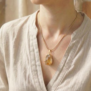 Puede incluir: Un collar de tono dorado con un colgante de citrino en bruto e irregular de aspecto amarillo y transl&uacute;cido. El colgante cuelga de una cadena fina y lisa de tono dorado que descansa sobre la clav&iacute;cula, mostrando la piedra natural.