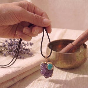 May include: A pendant necklace featuring a large, irregular purple amethyst stone accented with a smaller, round turquoise stone. The stones are set in a gold-toned metal frame and hang from a simple, dark brown cord necklace.
