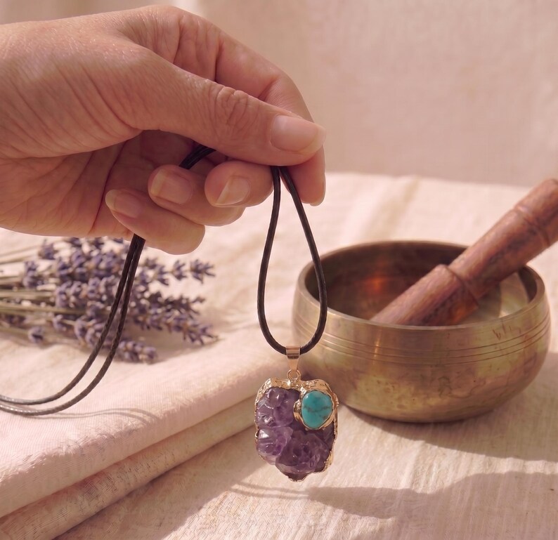 May include: A pendant necklace featuring a large, irregular purple amethyst stone accented with a smaller, round turquoise stone. The stones are set in a gold-toned metal frame and hang from a simple, dark brown cord necklace.