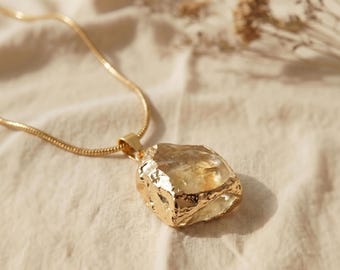Puede incluir: Un collar de cadena de serpiente dorada con un gran colgante de cristal amarillo translúcido e irregular. El cristal está engastado en una montura metálica texturizada de color oro que envuelve sus bordes dentados y en bruto.