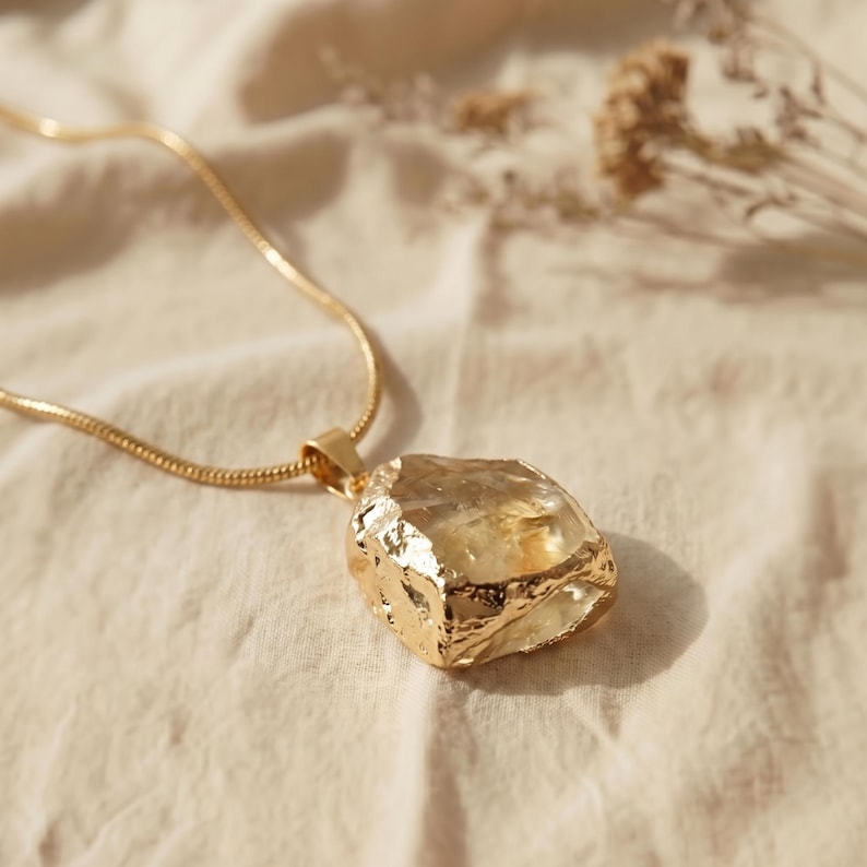 Puede incluir: Un collar de cadena de serpiente dorada con un gran colgante de cristal amarillo transl&uacute;cido e irregular. El cristal est&aacute; engastado en una montura met&aacute;lica texturizada de color oro que envuelve sus bordes dentados y en bruto.
