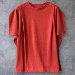 May include: A solid rust-orange t-shirt featuring a crew neckline and wide, short sleeves with a distinct layered or folded construction. The garment has a straight hemline and a relaxed fit, showcasing a plain, textured fabric throughout the design.