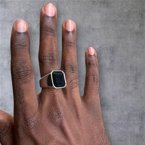 May include: A silver signet ring with a large, rectangular black stone. The ring is on a hand with a gray background. The ring's design is simple and modern, with a polished silver band.