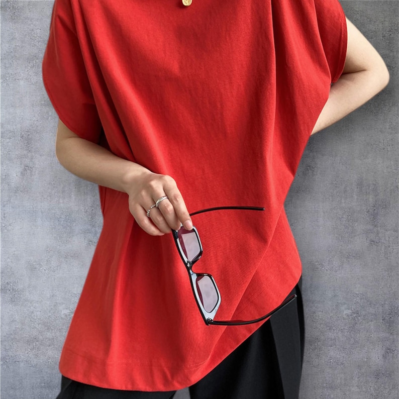 May include: A bright red short-sleeved top with a loose, relaxed fit and a high neckline. The garment features a solid colour design with wide sleeves and a straight hemline, paired with black trousers and black-framed sunglasses held in hand.