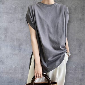 May include: A charcoal grey short-sleeved t-shirt with a crew neckline and a loose, relaxed fit. The shirt features side slits at the hem and is paired with plain white trousers. The fabric appears to be a solid, matte cotton jersey material.
