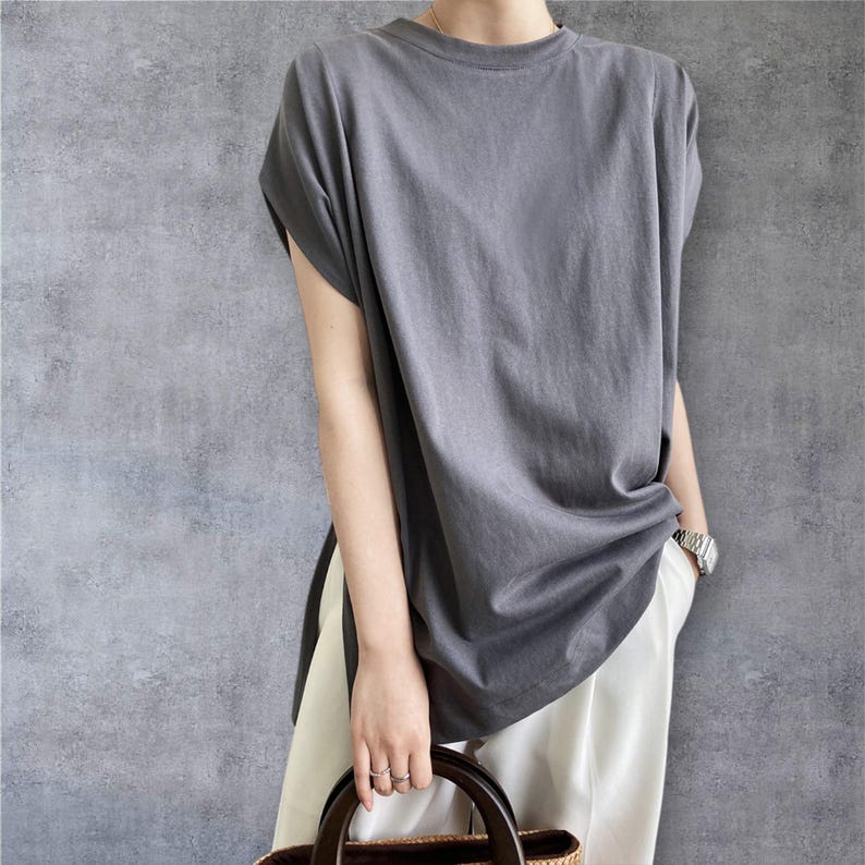 May include: A charcoal grey short-sleeved t-shirt with a crew neckline and a loose, relaxed fit. The shirt features side slits at the hem and is paired with plain white trousers. The fabric appears to be a solid, matte cotton jersey material.