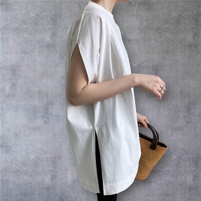 May include: A white sleeveless tunic top featuring a high crew neckline, wide arm openings, and deep side slits at the hem. The garment has a loose, relaxed fit and is paired with a small, structured brown woven handbag with dark handles.