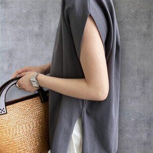 May include: A dark grey sleeveless top with wide arm openings and side slits, paired with a woven straw bag featuring dark brown leather handles. The top is worn over white fabric, and a silver-toned metal wristwatch is visible on the wrist.