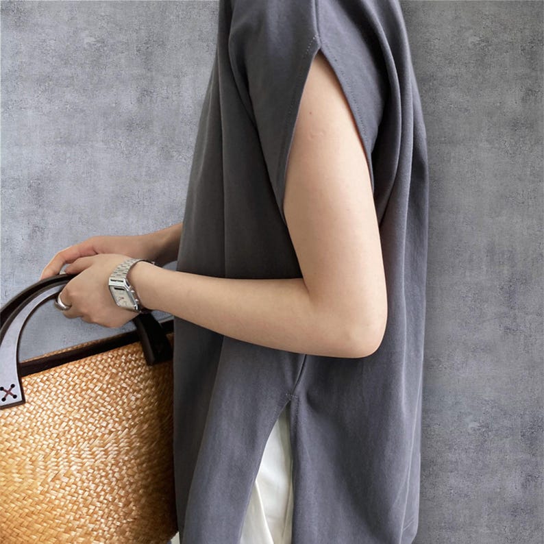 May include: A dark grey sleeveless top with wide arm openings and side slits, paired with a woven straw bag featuring dark brown leather handles. The top is worn over white fabric, and a silver-toned metal wristwatch is visible on the wrist.