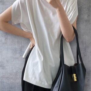 May include: A loose-fitting white short-sleeved t-shirt with a wide neckline and a black leather tote bag featuring a gold-toned metal clasp closure. The t-shirt has a relaxed silhouette and is paired with black trousers, creating a simple contrast.