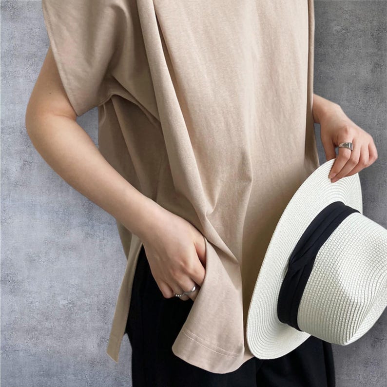 May include: A beige short-sleeved t-shirt with side slits and a loose fit, paired with black trousers and a white straw fedora hat featuring a wide black ribbon band. The t-shirt has a simple crew neckline and a smooth, solid-coloured fabric texture.