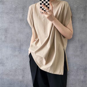 May include: A beige short-sleeved top with a crew neckline and a loose, relaxed fit. The garment features a straight hemline with deep side slits on both sides, extending from the bottom edge. The fabric appears to be a solid, smooth cotton blend.