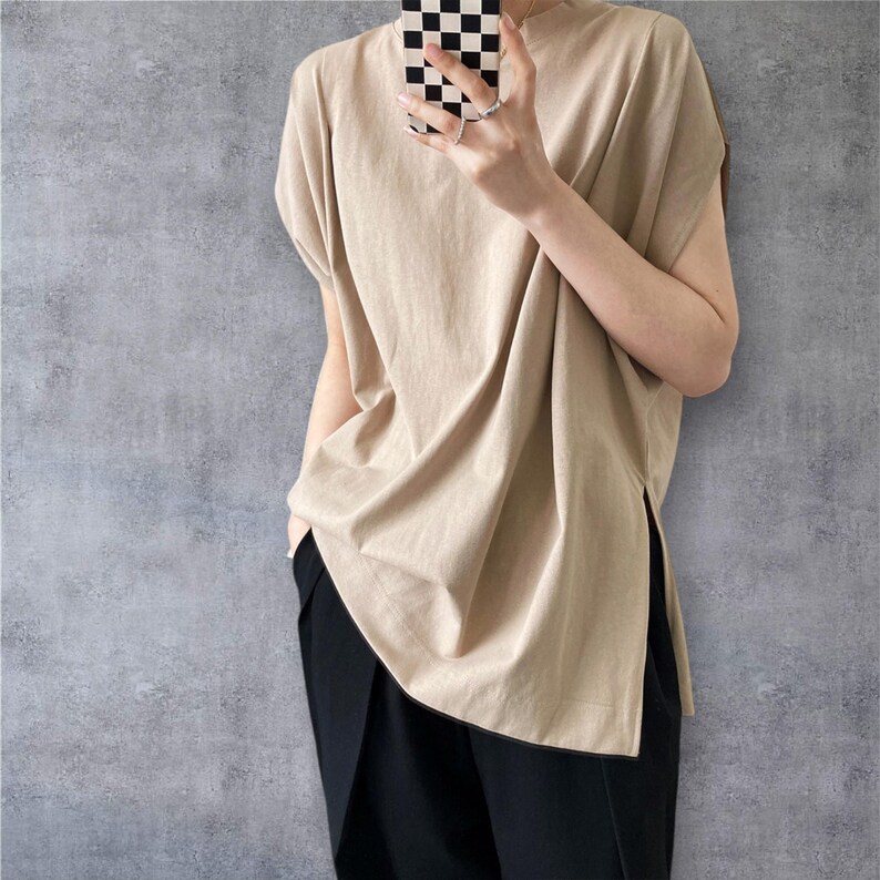 May include: A beige short-sleeved top with a crew neckline and a loose, relaxed fit. The garment features a straight hemline with deep side slits on both sides, extending from the bottom edge. The fabric appears to be a solid, smooth cotton blend.