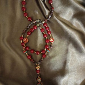 May include: A red beaded necklace with gold accents and a decorative pendant. The necklace features a series of red beads, interspersed with smaller beads and gold-colored spacers. The pendant has a turquoise and red design.