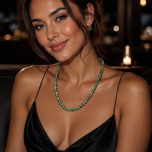 May include: A green beaded necklace with gold accents. The necklace is worn over a black satin dress. The image is a portrait of a person with dark hair and a warm complexion, wearing small gold hoop earrings.