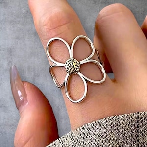 Könnte beinhalten: Ein silberner Blumenring mit einem detaillierten, strukturierten Mittelteil. Der Ring hat fünf offene Blütenblätter, die ein Blumenmuster bilden. Der Ring wird an einem Finger getragen und zeigt das Design und den Stil des Schmucks.